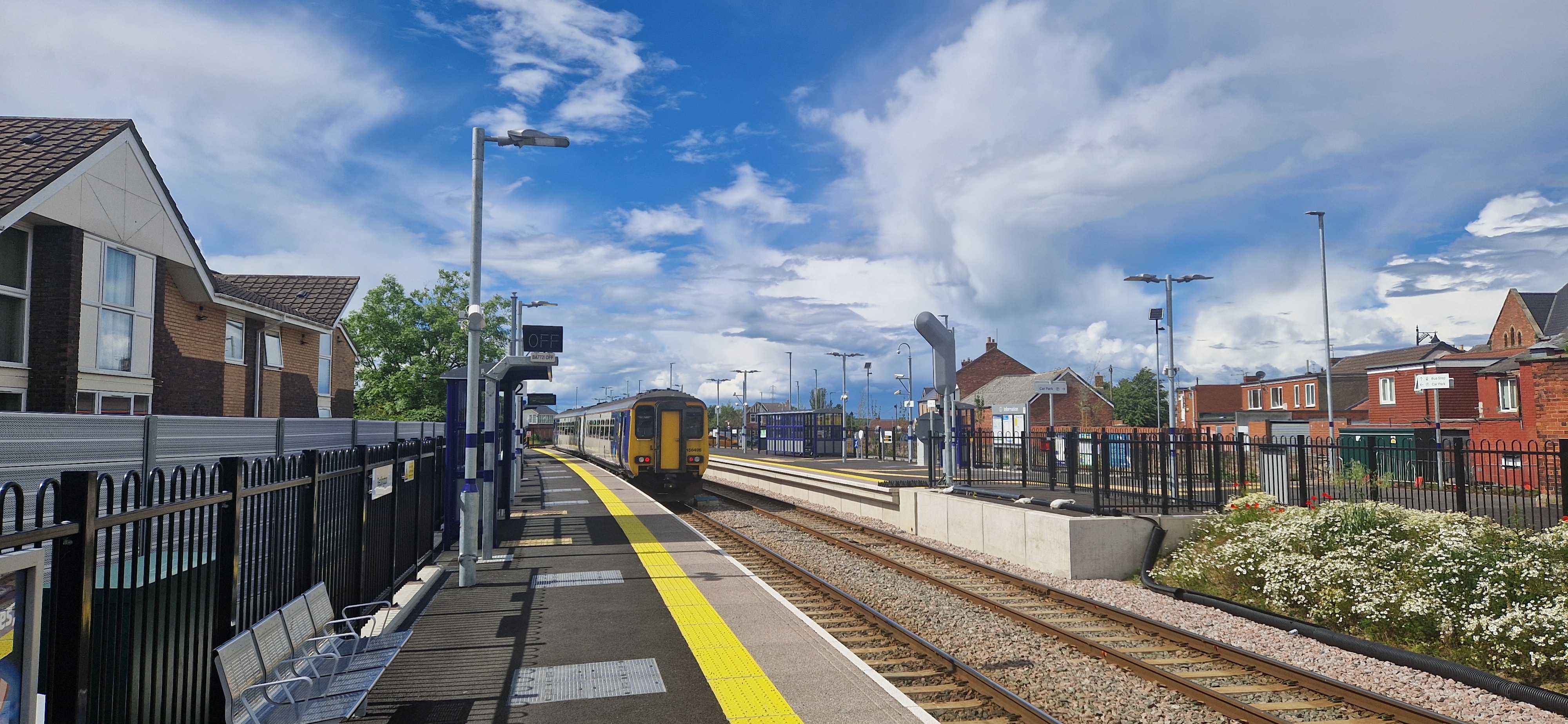 A Class 158 leaving the small, tidy provincial station in Bedlington, with a few amenities like benches and a covered waiting area, surrounded by exurban duplexes